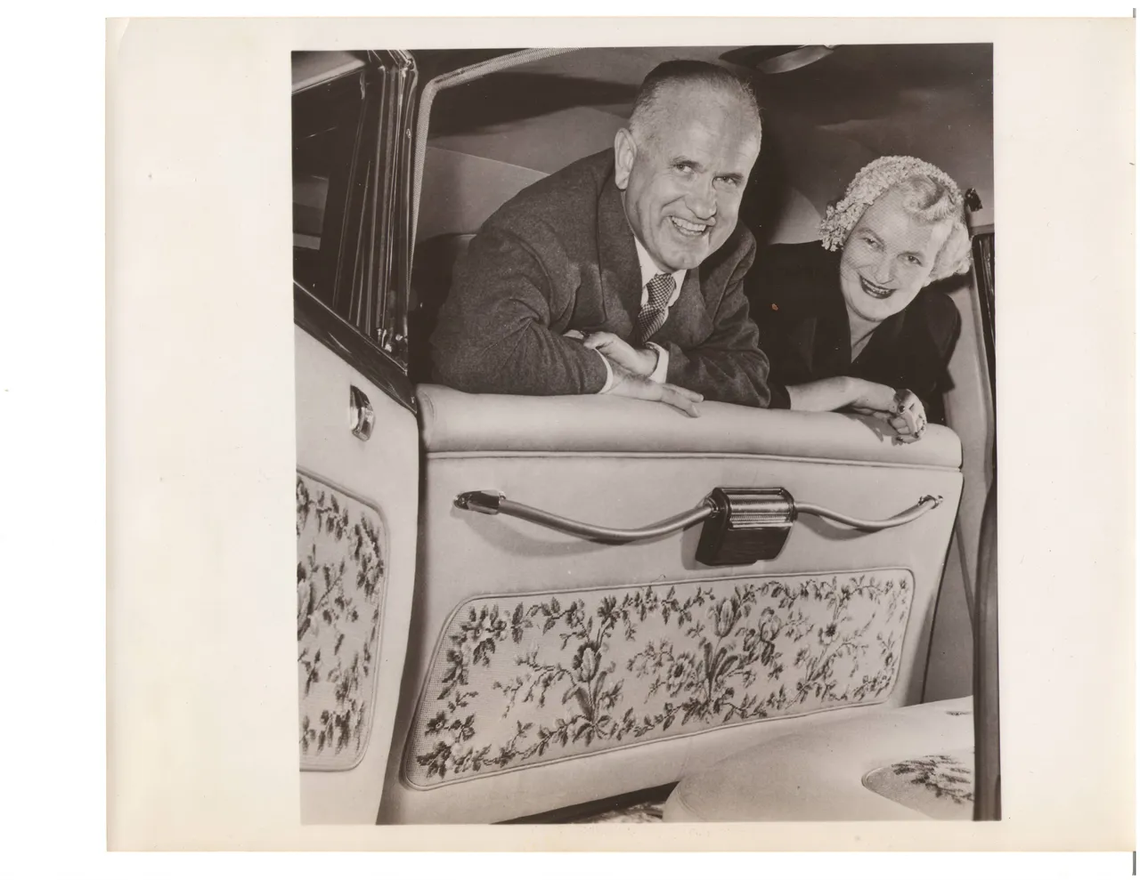 A woman and an older man inside a car whose door and seat panels are covered in floral needlepoint.